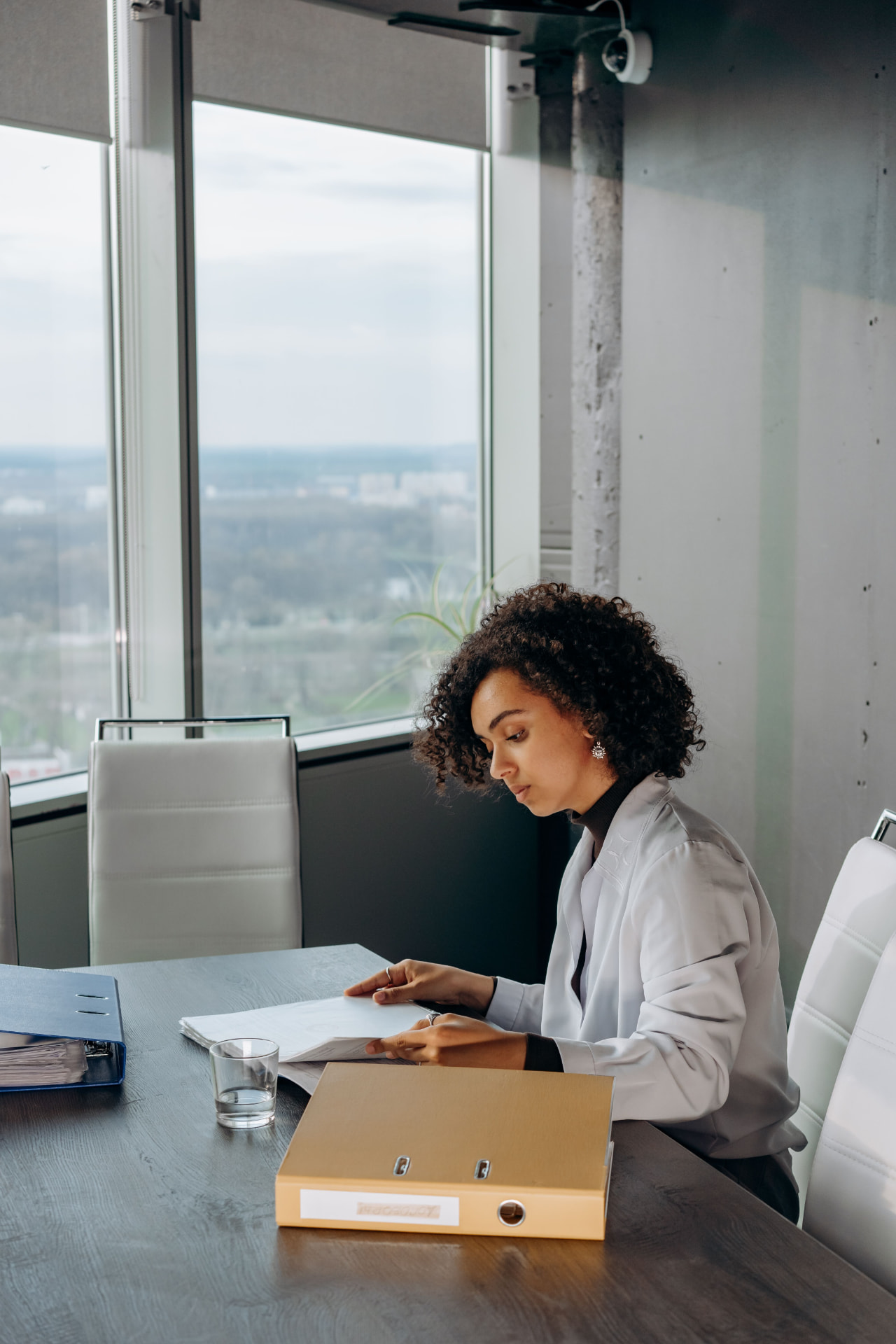 Young professional seated at a conference table reviewing a stack of documents, cityscape visible through the windows behind her