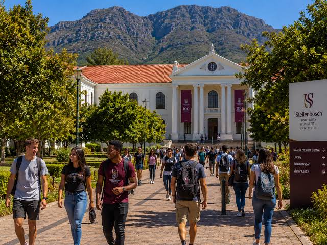 Students walking along a brick pathway toward the historic main building at Stellenbosch University, mountains rising behind
