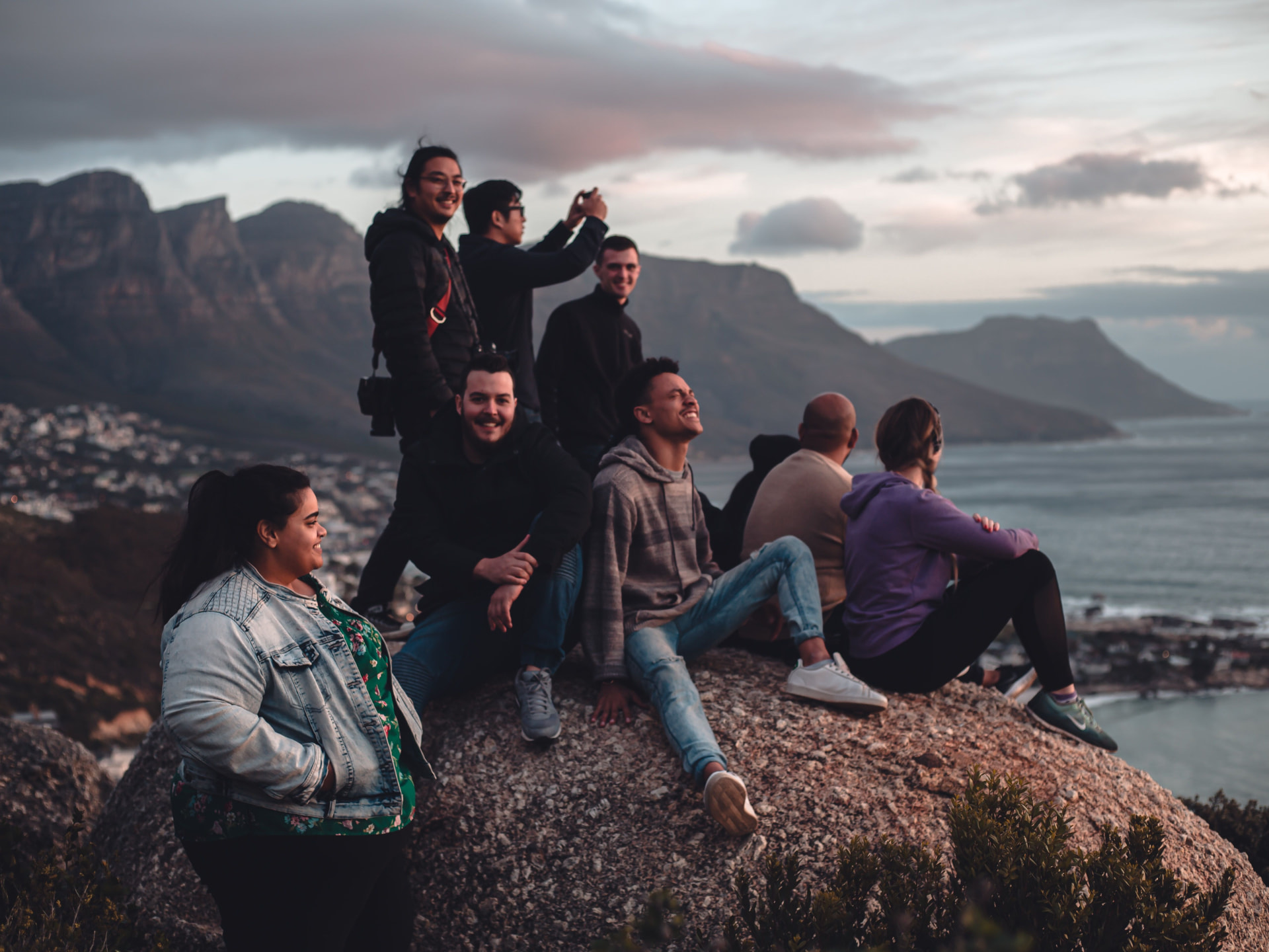Group of students gathered on a rocky mountain overlook at dusk, laughing and taking in the view of the ocean and mountain range